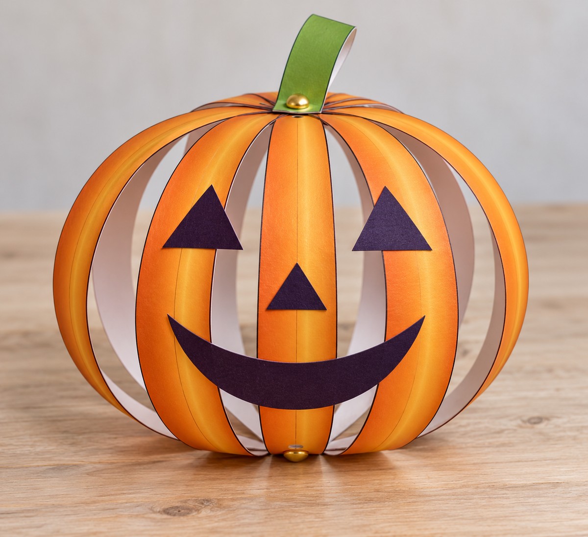 Illustrated assembly preview: eight shaded orange paper strips curve between top and bottom fasteners to form a hollow pumpkin, with a green stem and dark-purple face.